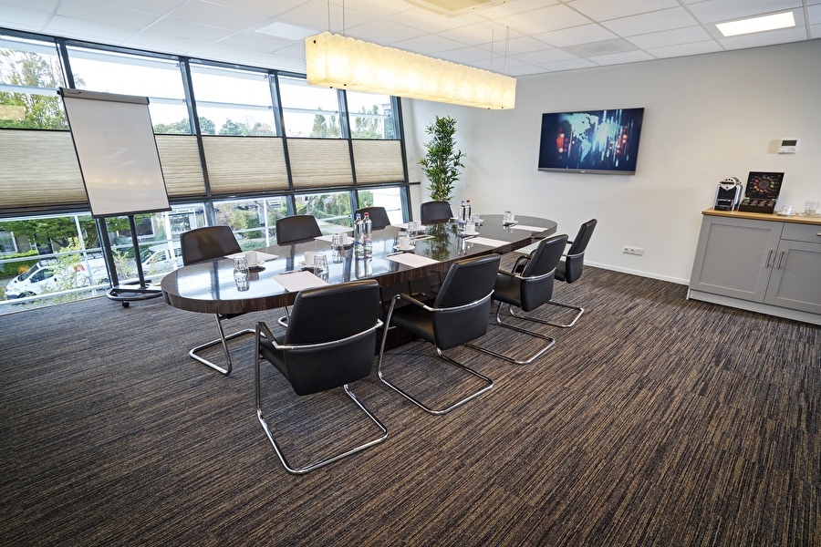 Moderne vergaderruimte in Amrâth Apart-Hotel Schiphol Badhoevedorp met ovale tafel, leren stoelen, grote ramen en een scherm aan de muur.