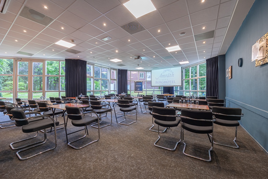 Vergaderzaal Amrâth Berghotel Amersfoort met houten tafels met daaromheen vier zwarte stoelen voor in de ruimte staat een centrale tafel met een beamer en een flipover naast een blauwe muur