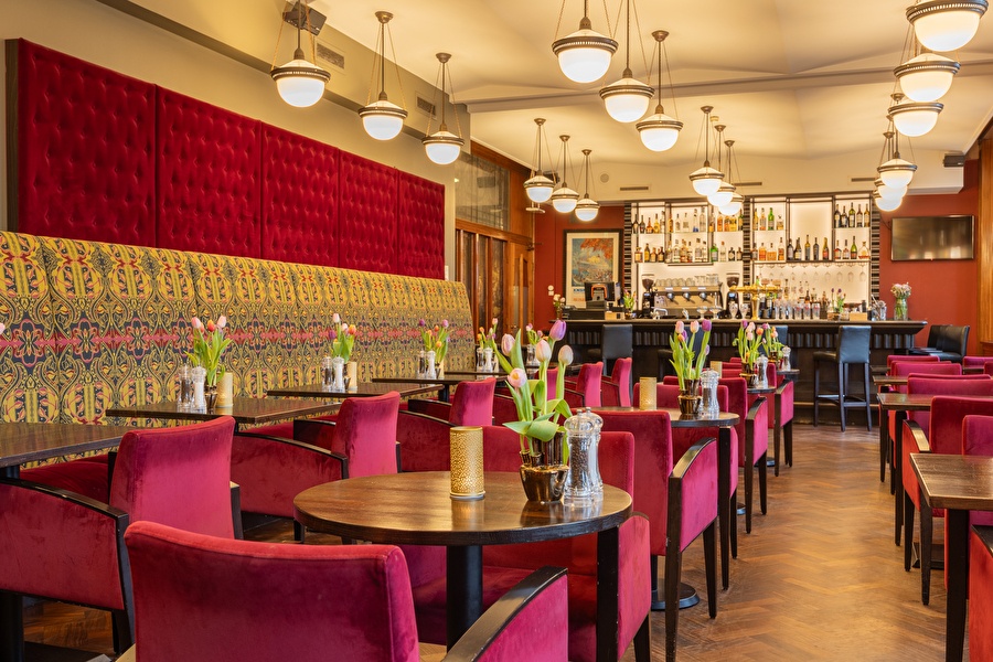 Elegante bar-lounge van Grand Hotel Amrâth Amsterdam met fluwelen rode stoelen, bloemen op tafel en bar op de achtergrond.