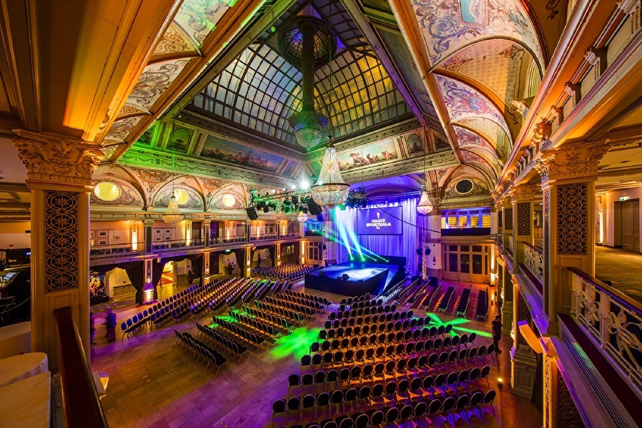 De sfeervol uitgelichte Kurzaal van Grand Hotel Amrâth Kurhaus met rijk versierde wanden en plafond, glazen koepel en podiumopstelling met rijen stoelen.