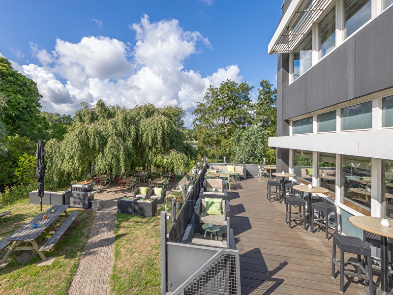 Lounge- en dinerterras aan het water bij Amrâth Airport Hotel Rotterdam, omgeven door bomen en natuur.