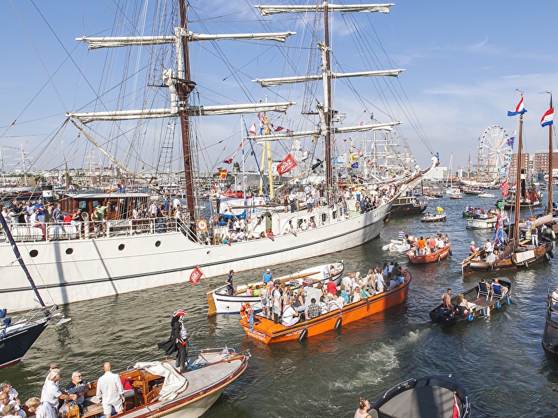 Drukke en feestelijke taferelen op het IJ tijdens SAIL Amsterdam, met tall ships, historische boten en veel mensen onder een stralende blauwe lucht.