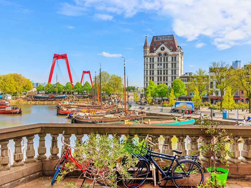 View of the Old Harbor in Rotterdam with historical ships, bicycles in the foreground, the White House, and the iconic red Willemsbrug under a blue sky.