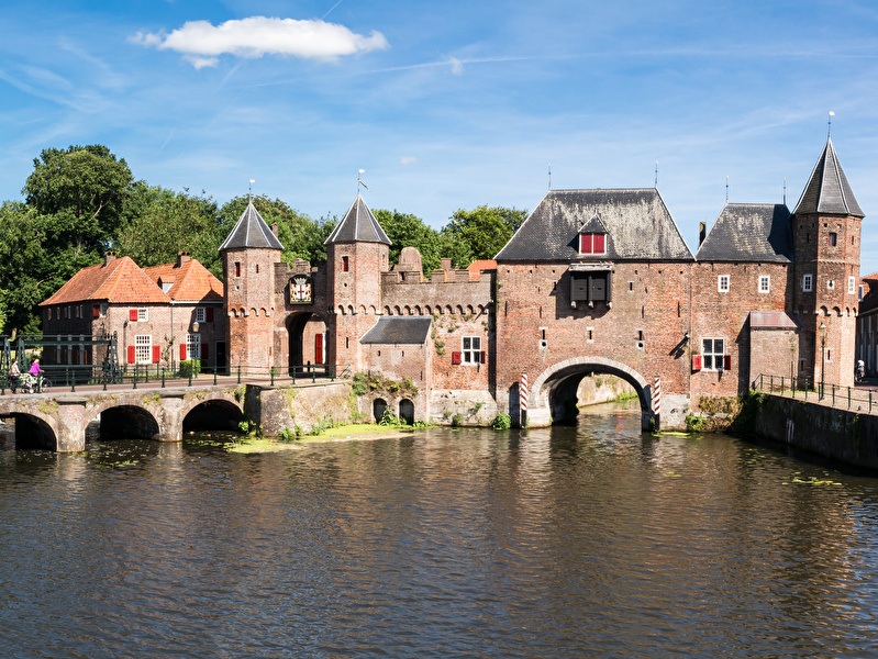 Medieval Koppelpoort in Amersfoort with a stone bridge over the water, surrounded by historic buildings and green trees on a sunny day.