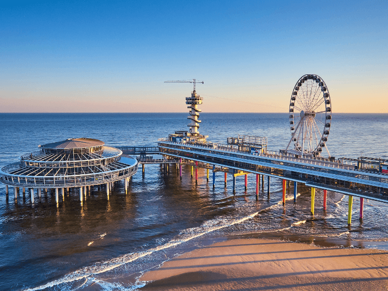 The Pier of Scheveningen with ferris wheel, tower, and pavilions above the sea, at sunset with a calm surf on the beach.