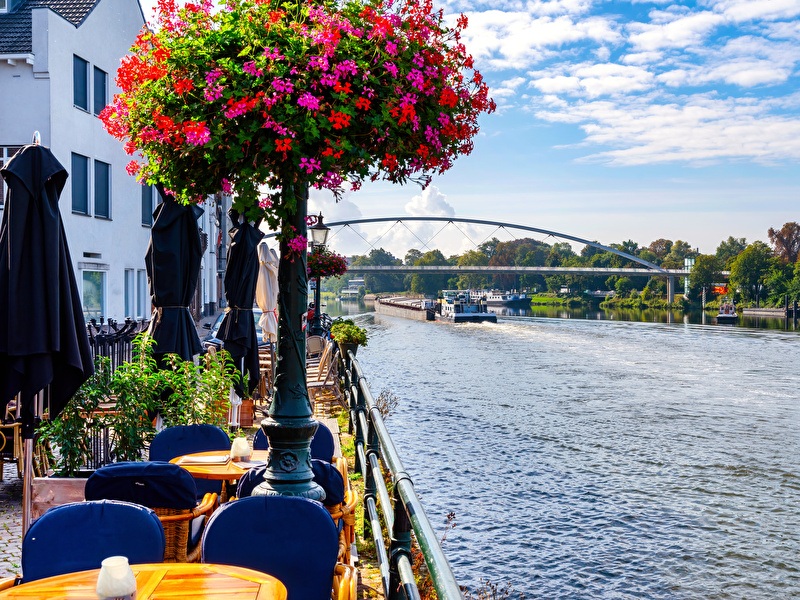 Terraces on the banks of the Maas in Maastricht with a view of a bridge and passing ships, surrounded by colorful flowers and green trees.