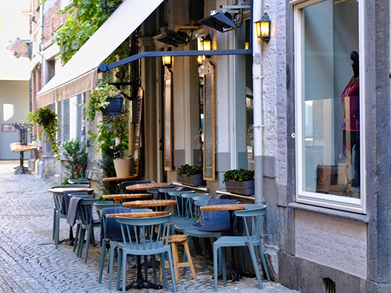 Cozy terrace in a narrow street in Maastricht with wooden tables, blue chairs and plants under a white awning by a characteristic building.