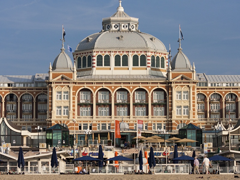  Imposante voorgevel van Grand Hotel Amrâth Kurhaus in Scheveningen, met klassieke koepel, torentjes en sierlijke boogconstructies, gezien vanaf het strand op een zonnige dag.