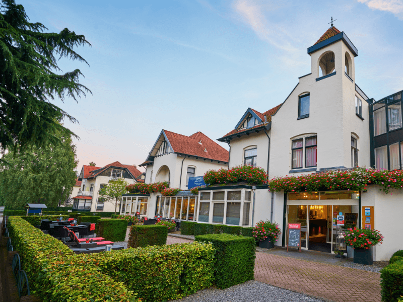 Vooraanzicht van Amrâth Hotel Media Park Hilversum met klassieke gevels, bloembakken vol kleurrijke bloemen en een zonnig terras omringd door verzorgde heggen.