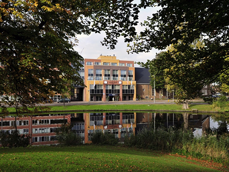 Vooraanzicht van Amrâth Hotel Alkmaar met moderne gevel en weerspiegeling in de vijver, omlijst door bomen.