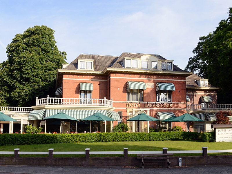 Klassieke gevel van Amrâth Hotel Lapershoek in Hilversum met groene luifels, terras met parasols en omliggende bomen.