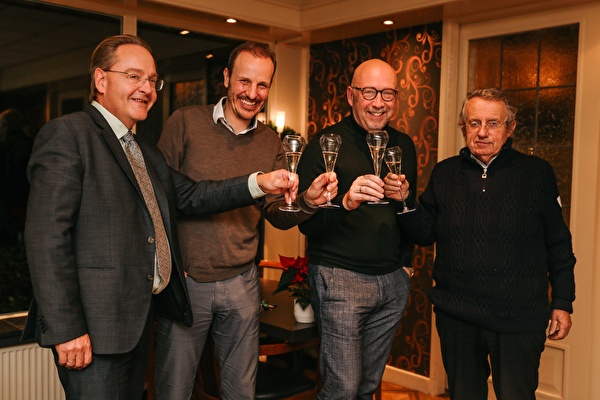 Vier mannen heffen lachend hun glazen tijdens een toost in een warme, sfeervol ingerichte kamer, waarschijnlijk ter gelegenheid van een zakelijke mijlpaal of viering.