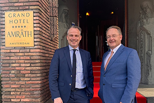 Twee mannen in pak poseren glimlachend voor de ingang van Grand Hotel Amrâth Amsterdam, naast een bord met de hotelnaam en voor een rode loper.