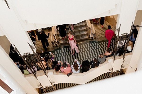 Een modeshow op een brede trap in een elegant art-nouveau-interieur, met modellen en een applaudisserend publiek aan weerszijden.